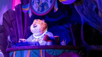 An animatronic Clawhauser wearing a purple Zoogether Day shirt and glow necklace, sitting behind a desk during a show of Zootopia: Better Zoogether