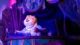 An animatronic Clawhauser wearing a purple Zoogether Day shirt and glow necklace, sitting behind a desk during a show of Zootopia: Better Zoogether